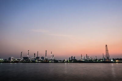 Illuminated factory by sea against sky during sunset