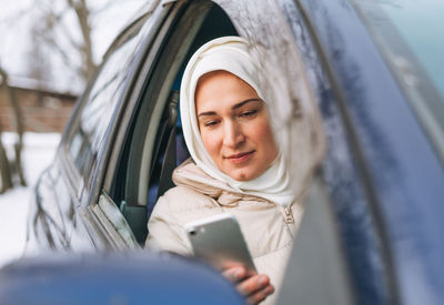 Beautiful smiling young muslim woman in headscarf in clothing using mobile in right-hand-drive car