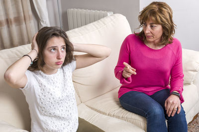 Daughter covering ears with hands while mother talking on sofa at home