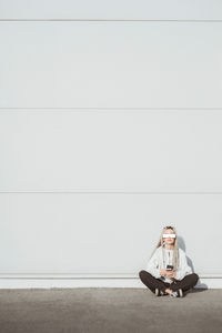 Full length of woman with dreadlocks holding mobile phone while sitting against wall