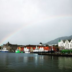 Rainbow over river by buildings in city against sky