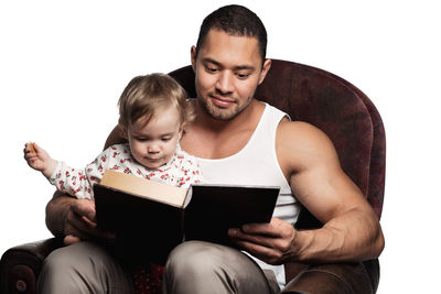 Father reading book with cute daughter while sitting on chair against white background