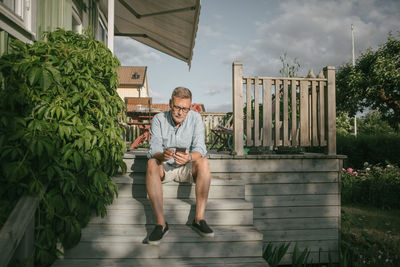 Full length of senior man using mobile phone while sitting on steps at porch