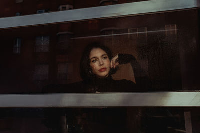 Through glass of thoughtful female standing near window and observing street while looking away