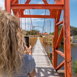 Rear view of a blonde woman taking a photo with smartphone of a pedestrian suspension bridge.