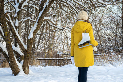 Young woman in yellow jacket with ice skates walking through snowy park