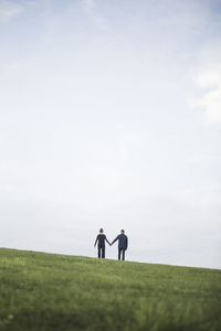 Rear view of couple holding hands standing on grassy field against cloudy sky