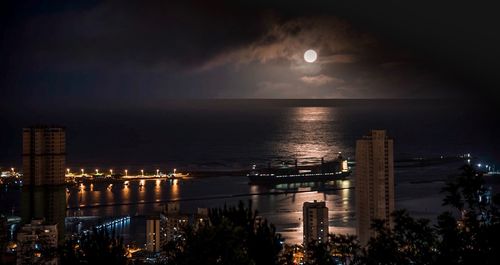 Illuminated city by sea against sky at night