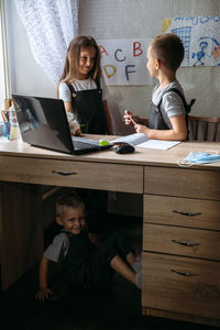 Cute boy hiding under table while sibling talking at home