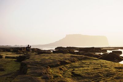 Mid distance view of man photographing on rocks at beach against sky