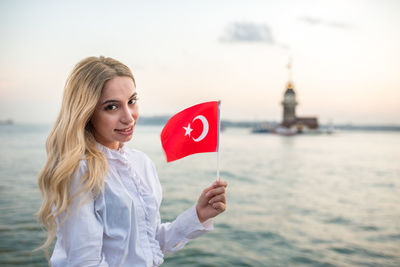 Portrait of smiling young woman holding turkish flag at beach against sky during sunset