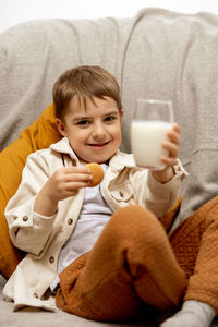 Little adorable boy sitting on the couch at home, drinking milk with cookie. fresh milk in glass