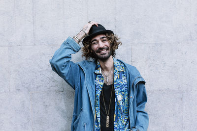 Smiling young man touching hat while standing in front of wall