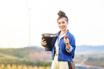 Portrait of smiling young woman holding strawberries against sky