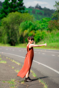 Side view of young woman wearing sunglasses hitchhiking while standing on road