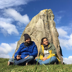 Couple sitting on grassy field against sky at avebury stone circle