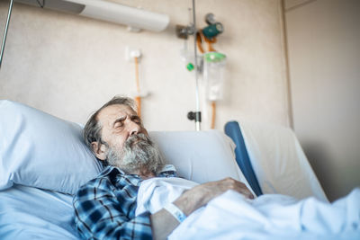 Calm aged man with beard lying under blanket on bed in hospital ward and sleeping