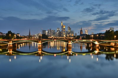 Illuminated bridge over river by buildings against sky at night