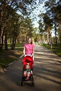 Portrait of happy mother pushing son in carriage amidst trees on footpath at park