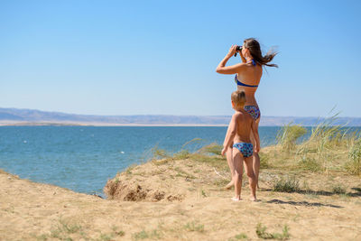 Woman looking through binoculars with son at beach against clear sky