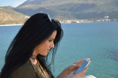 Side view of young woman using phone against sea