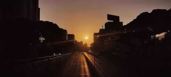 Road amidst silhouette buildings against sky during sunset