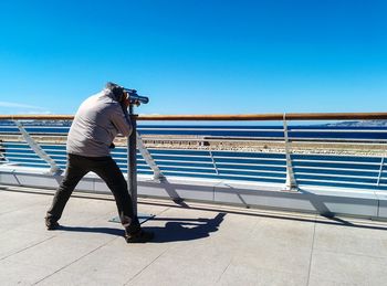 Full length rear view of man looking from coin-operated binocular against clear blue sky