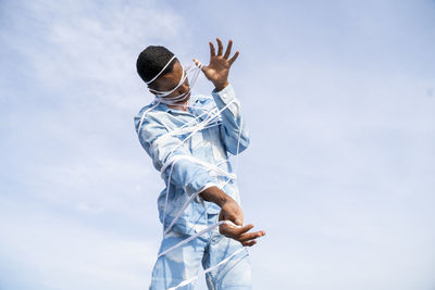 Young man wearing cloud patterned denim outfit struggling to get free from rope while standing against sky