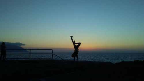 Silhouette man practicing handstand at beach against clear sky during sunset