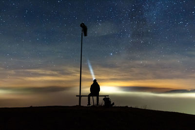 Person with flashlight sitting on bench against milky way