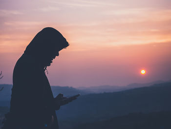 Silhouette of man enjoying the sunrise at mountain range and using smartphone.