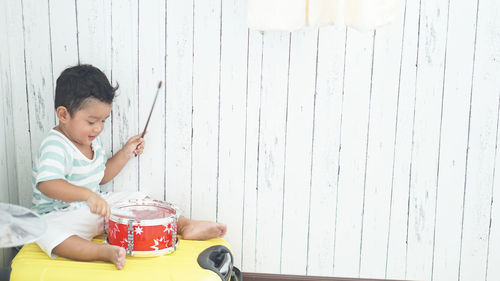 Full length of boy playing drum while sitting on suitcase against wooden wall