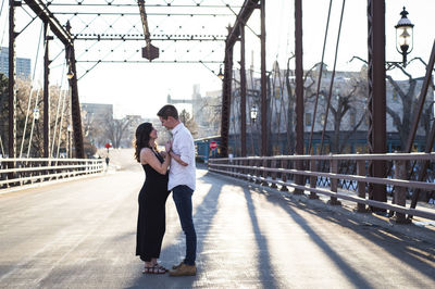 Side view of romantic couple looking at each other while standing on bridge against clear sky