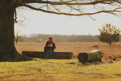 Full length of smiling mid adult woman sitting on tree trunk against sky during sunny day