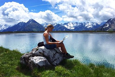 Side view of woman reading map while sitting on rock at riverbank against snowcapped mountain