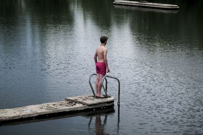 Rear view of shirtless boy standing on pier over lake