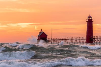 Waves splashing on pier and lighthouse against sky during sunset