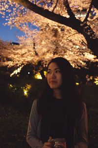 Portrait of young woman looking away at night with illuminated trees