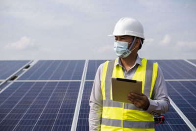 A young asian engineer wearing a helmet and white mask checks the cleanliness of solar panels. 