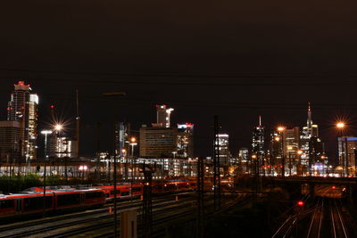 Illuminated railroad tracks amidst buildings against sky at night