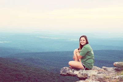 Portrait of smiling young woman sitting on cliff against sky