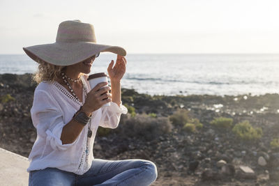 Smiling woman with disposable cup sitting at beach