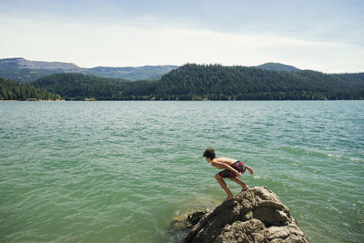 Boy bending on rock at lake against sky