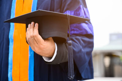 Midsection of man wearing graduation gown holding mortarboard