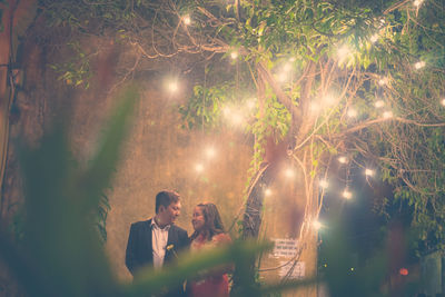 Couple standing below illuminated lighting decoration on tree at night