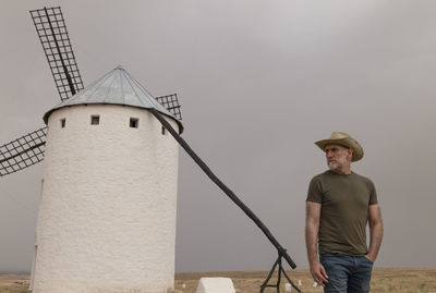 Full length of man with cowboy hat standing by spanish traditional white windmills against sky