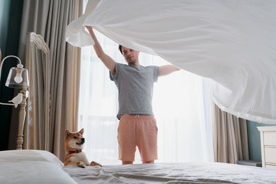 Young man and his funny dog are putting the bedding cover or mattress pad on the bed