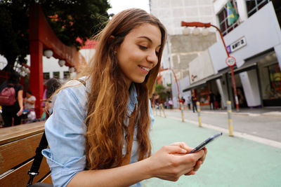 Smiling young woman using mobile phone against buildings in city