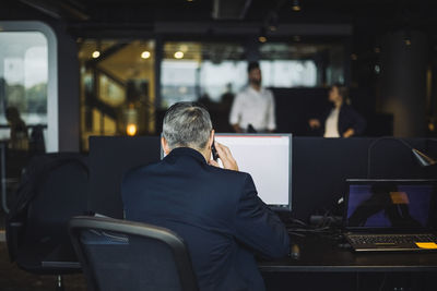 Rear view of businessman talking on smart phone while working on computer at office desk