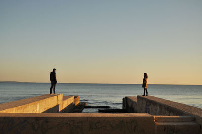 People standing on surrounding wall at beach against sky during sunset